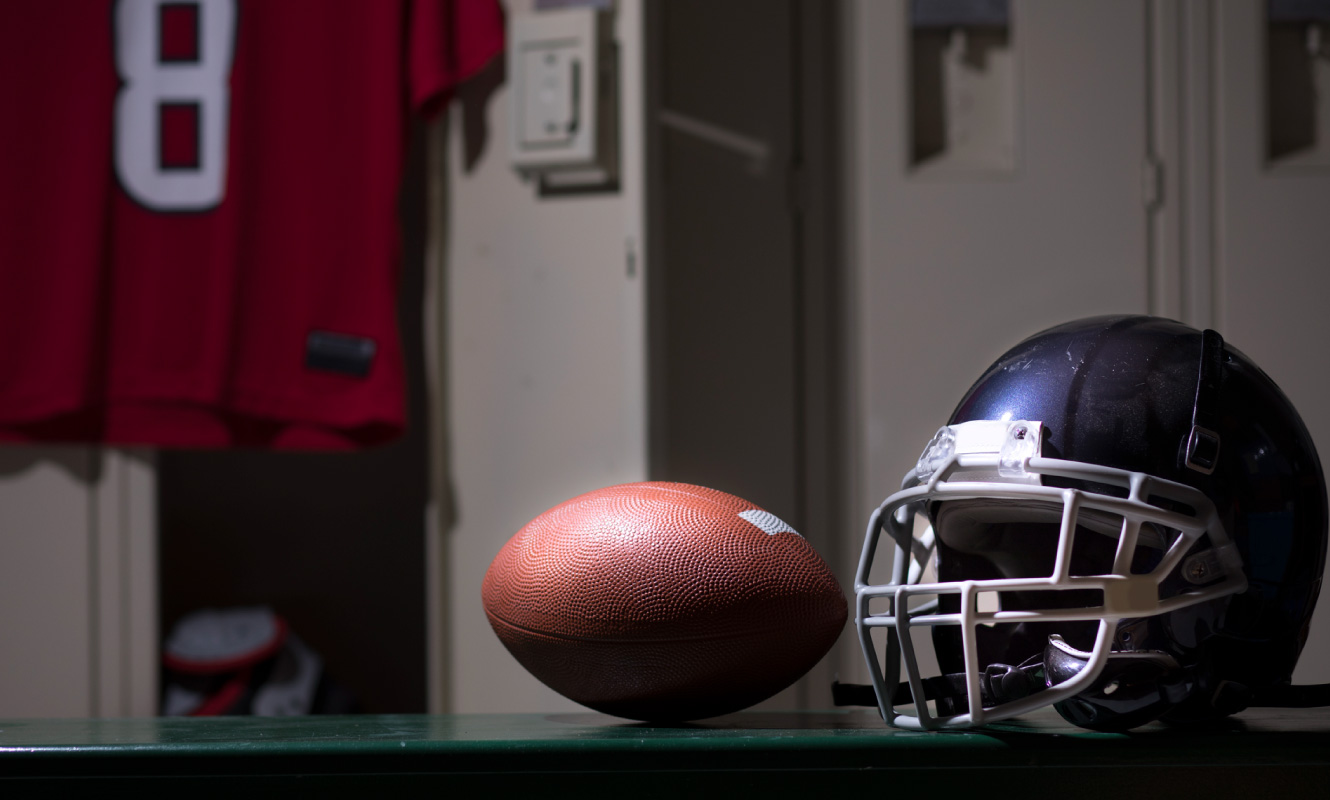 Close up of a football and helmet sitting on a bench in a locker room. There is a red jersey hanging in the background. 
