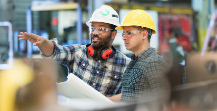 Two workers with hard hats, both are looking at something one man is pointing to off-camera.