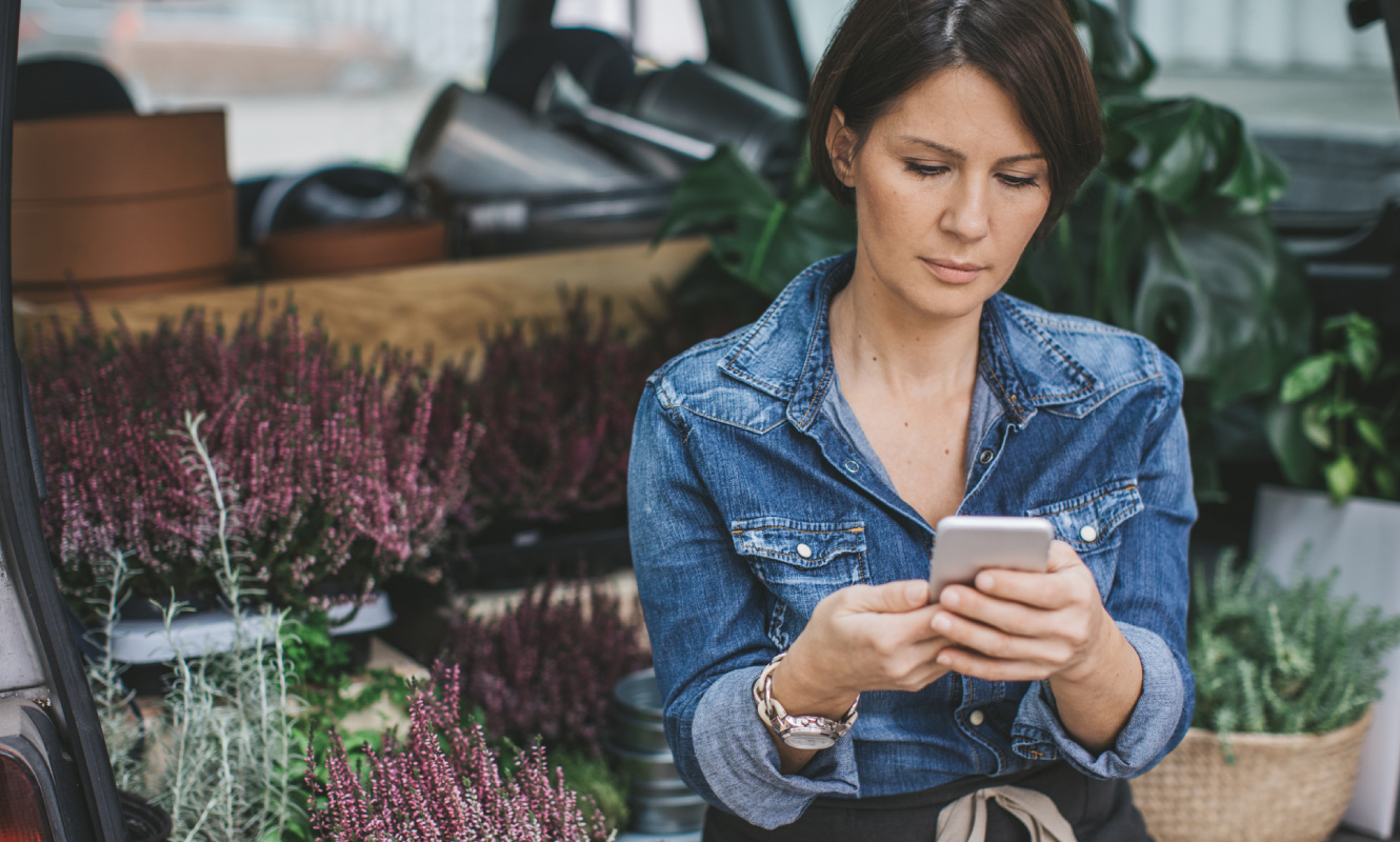 Woman looking at her smartphone. There is a car with various flowers and plants inside behind her.