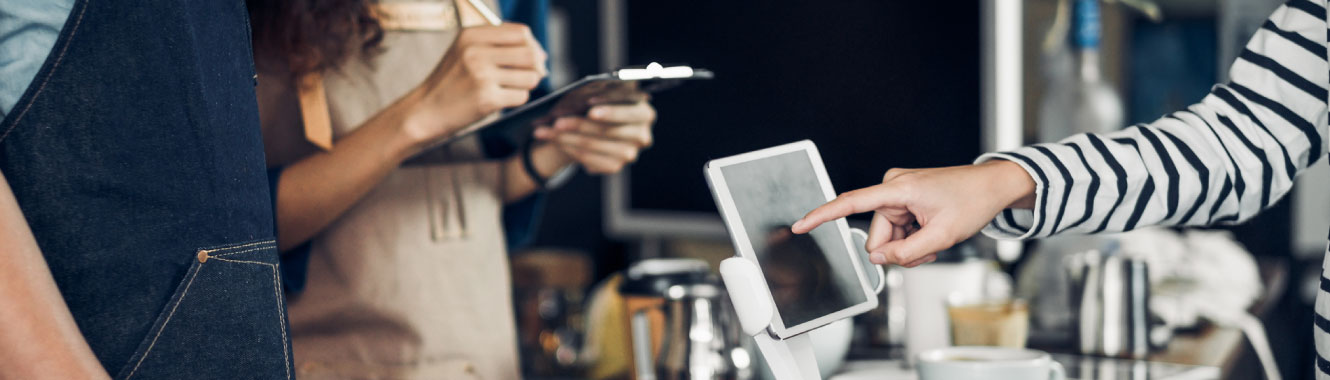 A person paying on a tablet screen at a coffee shop. Two people are behind the counter.