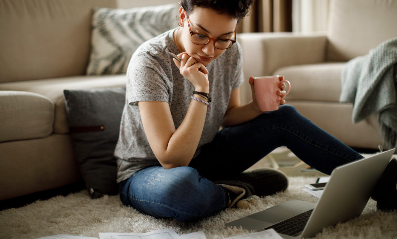 Young woman sitting in a living room looking down at a laptop that is on the floor.