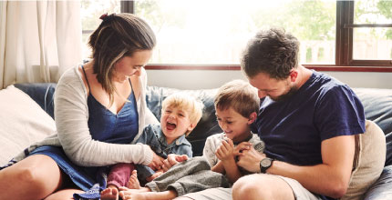 A mom and dad with two young kids laughing and playing inside on a couch.